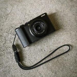 May include: Black digital camera with a lens that reads "FUJINON ASPHERICAL LENS SUPER EBC 1:1.8-5mm 1:2.8". The camera has a black wrist strap attached. The camera is on a grey concrete surface.