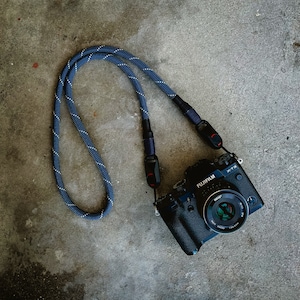 May include: A black camera with a blue and white camera strap. The camera has a lens with a silver ring and the text "FUJIFILM X-T3" printed on the top.