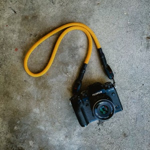 Pode incluir: Câmera Fujifilm X-T3 preta com uma alça de pescoço de corda amarela. A lente da câmera é visível e a alça está presa à lateral da câmera. A câmera está em uma superfície de concreto cinza.