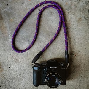 May include: A black camera with a purple and orange striped camera strap. The camera has the text "FUJIFILM" and "X-T3" on it.