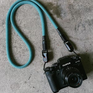 May include: A black Fujifilm X-T3 camera with a lens, suspended by a teal blue rope camera strap. The strap is made of woven material. The camera rests on a grey concrete surface. The camera strap is approximately 100 cm long.