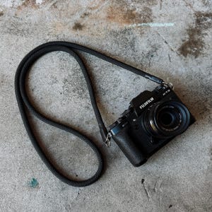 Puede incluir: Cámara Fujifilm X-T3 negra con lente negra y correa negra. La cámara está sobre una superficie de hormigón gris. La correa de la cámara es de material tejido.