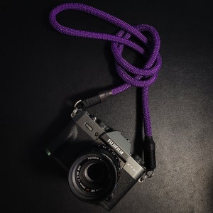 May include: A black camera with a purple camera strap. The camera has a lens with the text "FUJIFILM X-T30" and "FUJINON" printed on it.