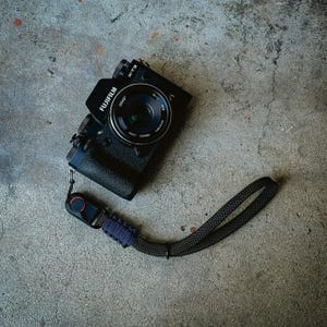 May include: A black Fujifilm X-T3 camera with a black lens and a black wrist strap. The camera is sitting on a grey concrete surface.