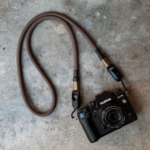 May include: A black Fujifilm X-T2 camera with a brown rope camera strap. The camera strap is made of a woven material and has black connectors. The camera lens is visible.