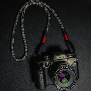 May include: A silver Canon EOS Elan II camera with a black Canon EF 50mm 1:1.4 lens attached. The camera has a black strap with red accents.