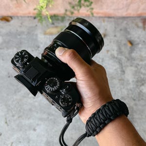 May include: Black digital camera with a black lens and a black wrist strap. The camera is held in a person's hand. The wrist strap is made of woven black material.