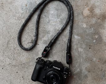 Könnte beinhalten: Schwarze Fujifilm X-T2 Kamera mit einem schwarz-grau gesprenkelten Kameragurt. Der Kameragurt ist mit schwarzen Anschlüssen befestigt. Das Kameraobjektiv ist zentriert. Die Kamera liegt auf einer grauen Betonoberfläche.