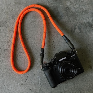 May include: Black Fujifilm X-T3 camera with a black lens and an orange rope camera strap. The strap has a black and gray pattern. The camera is on a gray concrete surface.