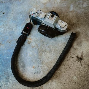 May include: A silver camera with a black lens and a black strap. The camera has multiple dials and buttons.