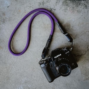 May include: A black Fujifilm X-T3 digital camera with a lens attached, featuring a purple braided rope camera strap. The strap is connected to the camera body using black plastic quick-release connectors and black heat-shrink tubing accents.