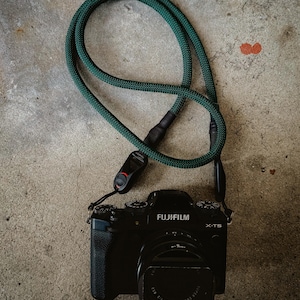 Puede incluir: Una cámara negra con una correa de cámara verde. La cámara tiene el texto "FUJIFILM" y "X-T5" en ella.