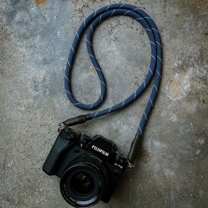 May include: A black camera with a blue and white camera strap. The camera has the text "FUJIFILM" and "X-T3" on it.