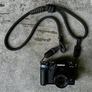 Könnte beinhalten: Schwarze Fujifilm X-T3 Kamera mit schwarzem Objektiv und schwarzem Kameragurt. Der Gurt besteht aus einem dicken, gewebten Material und hat verstellbare Schnallen. Die Kamera liegt auf einer grauen, strukturierten Oberfläche.