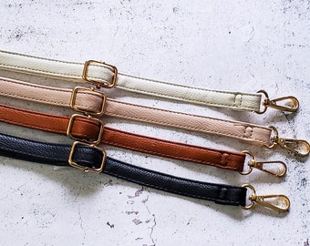 May include: Four leather bag straps in cream, tan, brown, and black, each with a gold-colored buckle and clasp. The straps are arranged parallel against a textured white background.