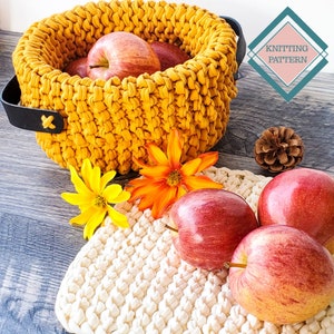 May include: A yellow crocheted bowl filled with red apples. The bowl has a black leather handle and is sitting on a wooden table. There is a white crocheted mat with more red apples on it. The image also includes a small pine cone and two flowers.