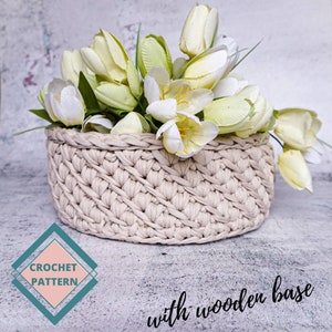 May include: A crocheted basket with a wooden base, filled with white and green artificial tulips. The basket is beige and has a woven pattern. The text "CROCHET PATTERN" is visible in the lower left corner of the image. The text "with wooden base" is visible in the lower right corner of the image.