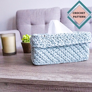 May include: A light blue crocheted tissue box cover with a woven pattern. The cover is sitting on a wooden table with a candle and a small plant in the background. The text "CROCHET PATTERN" is visible in the upper right corner of the image.