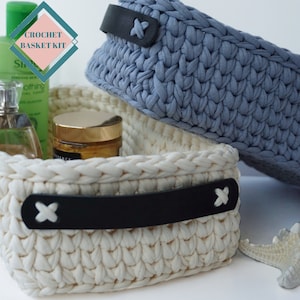 May include: Two crocheted baskets, one white and one blue, with black leather handles. The baskets are filled with various items, including a bottle of lotion, a jar of cream, and a small bottle of perfume. The text "CROCHET BASKET KIT" is visible in the top left corner of the image.