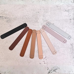 May include: Eight leather straps in various shades of brown, black, and gray, arranged in a semi-circle on a white textured surface. Each strap has multiple evenly spaced holes.