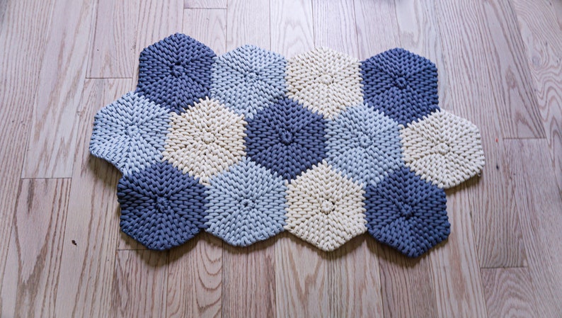 Puede incluir: Una alfombra tejida con cuerda gruesa en tonos azules y crema. La alfombra tiene forma de panal de abejas con secciones hexagonales.