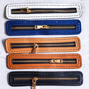 May include: Five zipper pulls with different colored leather straps. The straps are white, blue, brown, tan, and black. Each strap has a gold zipper pull and a row of small holes along the edges.