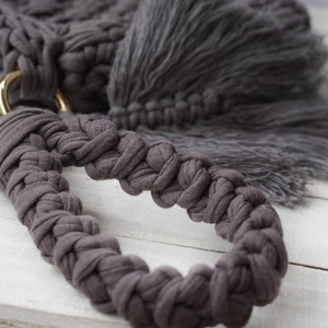 May include: A close-up of a gray crocheted bag handle with a gold metal ring. The handle is braided and has a tassel on the end.