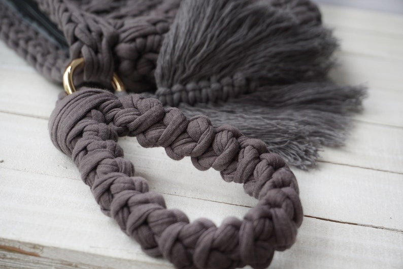 May include: A close-up of a gray crocheted bag handle with a gold metal ring. The handle is braided and has a tassel on the end.