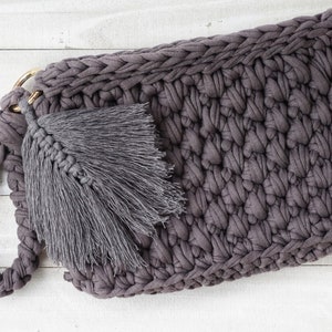 May include: A gray crocheted clutch bag with a tassel. The bag has a braided handle and a gold metal ring.