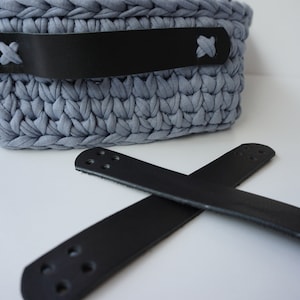 Puede incluir: Una cesta de crochet gris con asas de cuero negro. Las asas tienen detalles de costura y están unidas a la cesta con remaches metálicos.