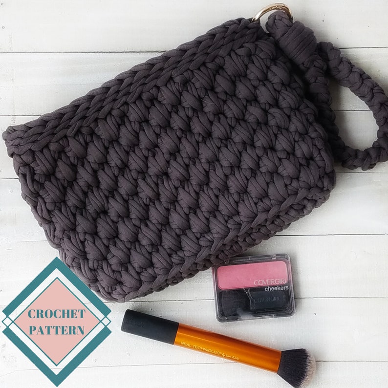 May include: A dark gray crocheted bag with a top handle. The bag is lying on a white surface with a pink blush compact and a makeup brush.