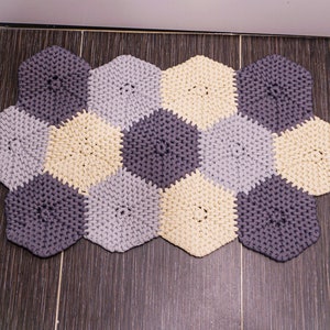 Puede incluir: Una alfombra tejida a crochet hecha de hilo gris, gris claro y crema. La alfombra tiene forma de panal y est&aacute; compuesta por formas hexagonales individuales.