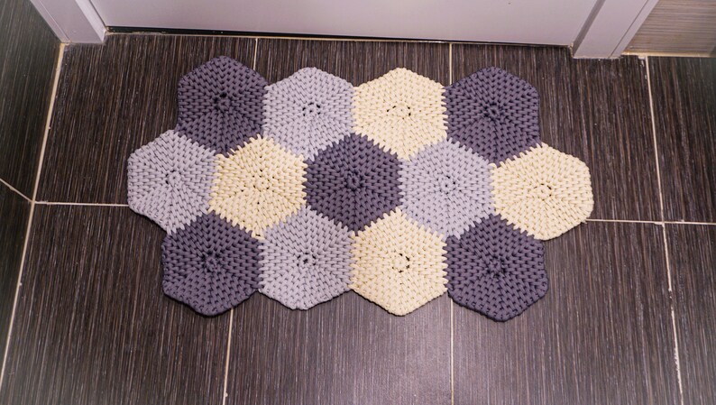 Puede incluir: Una alfombra tejida a crochet hecha de hilo gris, gris claro y crema. La alfombra tiene forma de panal y est&aacute; compuesta por formas hexagonales individuales.