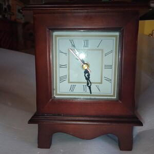 May include: A rectangular wooden clock with a dark brown finish. The clock face is white with black Roman numerals and black hands. The clock has a small, decorative top and sits on four small feet.