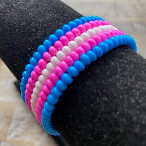 May include: A beaded ring in the colors of the transgender pride flag: blue, pink, white, pink, and blue. The ring is made of small, round beads and is displayed on a black surface.