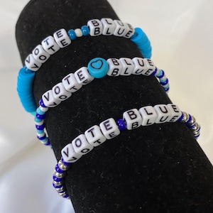 May include: Three beaded bracelets with the words "VOTE BLUE" spelled out in white letter beads. The bracelets feature blue and silver beads, with a heart-shaped bead on one. The bracelets are displayed on a black surface.
