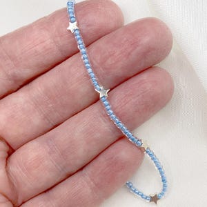 May include: A delicate beaded bracelet featuring small, light blue beads and silver-colored star charms. The bracelet is strung on a thin cord and is shown against a white background. The stars are evenly spaced along the bracelet.
