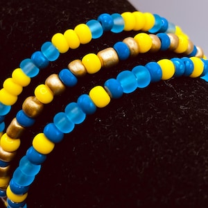 Puede incluir: Tres pulseras de cuentas con una mezcla de cuentas amarillas, azules y doradas. Las pulseras están hechas de pequeñas cuentas redondas ensartadas, creando un efecto en capas. Los colores son vibrantes y el diseño es sencillo.