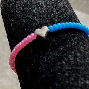 May include: A beaded bracelet featuring a single silver-toned heart charm positioned between a section of vibrant pink beads and a section of bright blue beads. The small, round, uniform beads are strung together to form a continuous circular band.