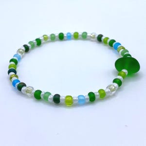 May include: A beaded bracelet featuring a variety of green, blue, and clear beads. The bracelet includes a large, flat, green glass bead. The beads are strung on a clear elastic cord.