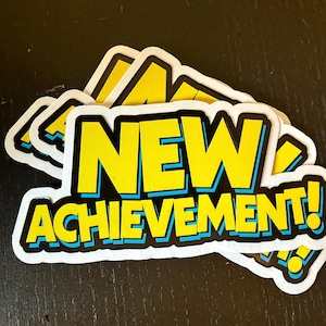 Puede incluir: Un montón de pegatinas con las palabras "NEW ACHIEVEMENT!" en letras amarillas con un contorno azul. Las pegatinas tienen un borde blanco y están sobre una superficie oscura.