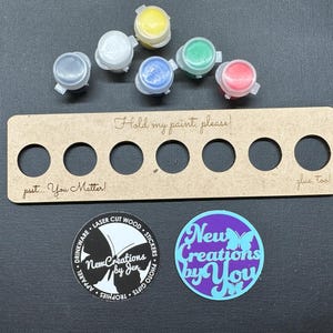 Może przedstawiać: Drewniany uchwyt na farbę z sześcioma okrągłymi otworami, mieszczący sześć małych słoiczków z farbą w różnych kolorach. Uchwyt zawiera tekst "Hold my paint, please!" i "put... You Matter!" Obecne są również dwie okrągłe naklejki.