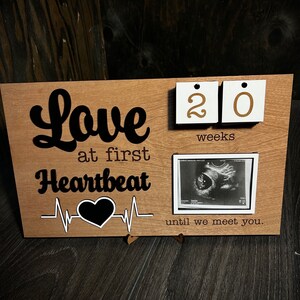 May include: A wooden sign with the text "Love at first Heartbeat" and a heartbeat graphic. The sign also has a small white frame with an ultrasound image and a calendar with the numbers "20" and the word "weeks". The text "until we meet you." is below the ultrasound image.