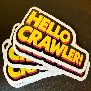 Puede incluir: Un montón de pegatinas con el texto "HELLO CRAWLER!" en amarillo, contorneado en rosa y negro, sobre un fondo blanco. Las pegatinas se superponen, con una superficie negra de fondo.