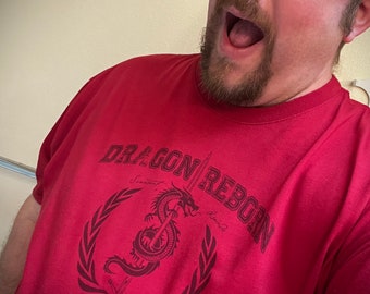 Puede incluir: Una camiseta roja con un gráfico en blanco y negro de un dragón con una espada. El texto "Dragon Reborn Academy" está impreso en la camiseta.