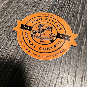 May include: Orange and black round sticker with the text "Two Rivers Animal Control" and the slogan "We don't need no bloody badgers!"  The sticker features a cartoon badger.