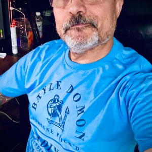 Può includere: Un uomo indossa una maglietta blu chiaro con un design di ancora blu scuro e il testo "Bayle Domon Fortune Pick'd Trading Co. Founded in Lillian Established ... See Pass..."