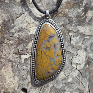 May include: A silver pendant with a yellow and white agate stone set in a beaded border. The pendant is hanging from a black cord.
