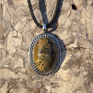 May include: Oval pendant necklace with a silver-colored bezel and a patterned stone in shades of green, brown, and yellow. The pendant hangs from a black braided cord.