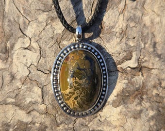 May include: Oval pendant necklace with a silver-colored bezel and a patterned stone in shades of green, brown, and yellow. The pendant hangs from a black braided cord.